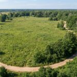 Na tym obrazie widać widok z lotu ptaka (z drona) na rozległą, niezabudowaną działkę budowlaną otoczoną przyrodą